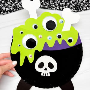 May include: A Halloween-themed cauldron decoration. The black cauldron has a skull on the front, green bubbling liquid, and large googly eyes. White bone accents and purple trim complete the design. The background features a skull and crossbones pattern.