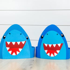 Op de afbeelding: Twee blauwe haai-vormige feesthoeden met witte tanden en rode monden. De hoeden hebben zwarte ogen en zijn gemaakt van gelaagd papier. De achtergrond is een witte houten muur en een lichte houten vloer.