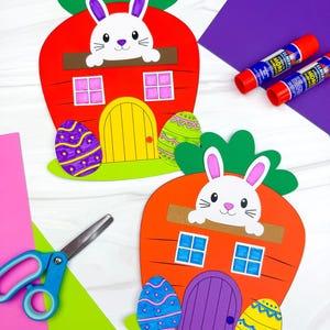 May include: Two carrot-shaped Easter crafts with a white bunny peeking out. The orange carrots have green tops, yellow and purple doors, and blue windows. Decorated Easter eggs are placed near the doors. Scissors, glue sticks, and colorful paper are also visible.