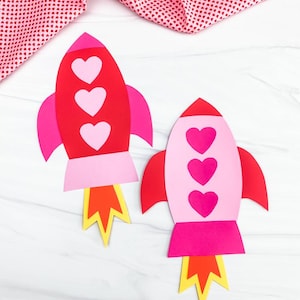 May include: Two colorful paper rockets, one red and one pink, each adorned with three heart-shaped cutouts. The rockets have pink fins and yellow and orange flames, perfect for Valentine's Day or a space-themed craft.