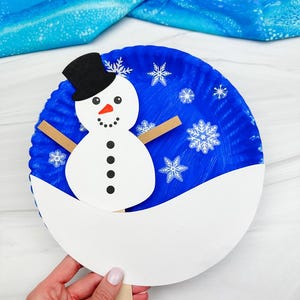 May include: A handmade snowman craft made from a blue paper plate, white paper, and a popsicle stick. The snowman has a black top hat, a carrot nose, and black button eyes and mouth. White snowflakes are scattered on the blue background.
