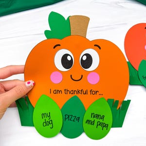 Op de afbeelding: Een papieren pompoenknutselwerk met een lachend gezicht, roze wangen en een bruine steel. De pompoen bevat de tekst "I am thankful for..." boven drie groene bladvormige uitsparingen met de handgeschreven woorden "my dog", "pizza" en "nana and papa".