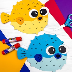 May include: Two paper pufferfish crafts, one yellow and one blue, featuring large white and black eyes, small fins, and V-shaped markings. The crafts are displayed alongside two red glue sticks and a pair of blue scissors on a white surface.
