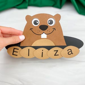 May include: A brown paper groundhog with a smiling face, large eyes, and the name "Eliza" below. The groundhog is made of brown and black paper, with a white tooth. The name is printed on brown ovals.