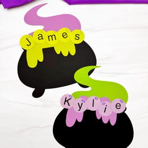 May include: Two black paper cauldrons with colourful cutouts. One cauldron features a purple steam plume and yellow circles spelling James. The other cauldron has a lime green steam plume and light purple circles spelling the name Kylie.