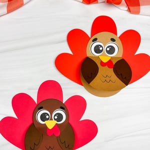 Puede incluir: Dos decoraciones de pavo hechas a mano. Cada pavo tiene un cuerpo marrón, plumas de cola rojas y una carúncula roja. Los pavos están hechos de papel y están colocados sobre un fondo blanco con un paño a cuadros rojo y blanco.