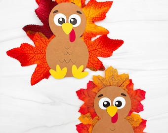 Puede incluir: Dos decoraciones de pavo hechas a mano. Cada pavo está hecho de papel marrón con un pico amarillo, una carúncula roja y patas amarillas. Los pavos tienen ojos grandes y están rodeados de hojas de otoño rojas, naranjas y amarillas. Un paño verde está en el fondo.