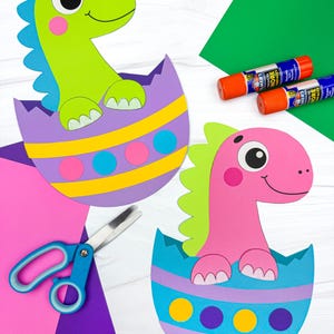May include: Two colorful paper dinosaur crafts emerging from decorated eggshells. One is green with blue accents, the other is pink with green accents. A pair of scissors and glue sticks are also visible.