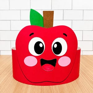 May include: A red paper apple headband featuring a cartoon face with large white eyes, black pupils, pink cheeks, and an open mouth with a red tongue. A green leaf and a brown stem are attached to the top. The text "simple everyday mom" is visible.