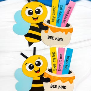 Op de afbeelding: Twee papieren bijenuitsparingen met zwart-gele strepen, blauwe vleugels en lachende gezichten. Elke bij houdt een pot honing vast met de tekst "BEE KIND" en kleurrijke stroken met positieve boodschappen zoals "Help anderen" en "Deel mijn speelgoed".