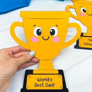 Op de afbeelding: Een gele trofee met een lachend gezicht en de tekst "World's Best Dad!" op een zwarte basis. De trofee heeft twee handvatten en roze wangen. Een tweede trofee is zichtbaar op de achtergrond.