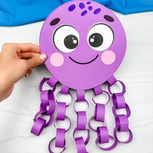 May include: A purple paper octopus craft with a smiling face, large eyes, and pink cheeks. The octopus has paper chain tentacles in a matching purple color. The background includes a blue fabric and a white surface.
