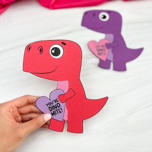 Op de afbeelding: Twee dinosaurussen van papier, één rood en één paars, elk met een hartvormig bord. De rode dinosaurus houdt een paars hart vast met de tekst "YOU'RE DINO MITE!". De achtergrond is roze. Valentijnsdag decoratie.