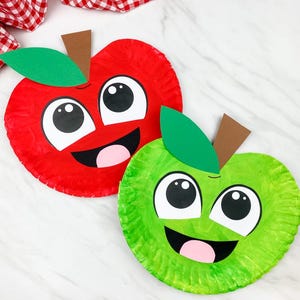 Op de afbeelding: Twee lachende appelvormige knutselwerkjes gemaakt van papieren borden. Eén is rood, de andere groen, elk met grote witte ogen, een roze tong en een bruine steel met een groen blad. Op de achtergrond een rood-witte geruite doek.