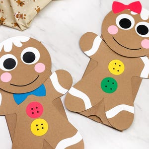 May include: Two brown paper gingerbread man-shaped gift bags with white icing details, black eyes, and pink cheeks. One has a blue bow tie and red and yellow buttons. The other has a red bow and green and yellow buttons. A beige fabric with gingerbread man prints is in the background.