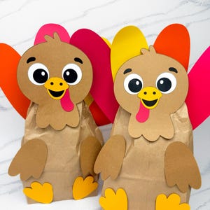 May include: Two handmade turkey-themed paper bag crafts. Each turkey has a brown paper bag body, with a cartoon turkey face, colorful feathers, and yellow feet. The turkey faces have large eyes, a yellow beak, and a red tongue.