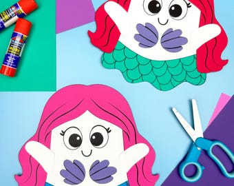 May include: Two mermaid paper crafts with red and pink hair, white faces, and blue and green tails. The mermaids have large eyes and are surrounded by craft supplies, including glue sticks and scissors.
