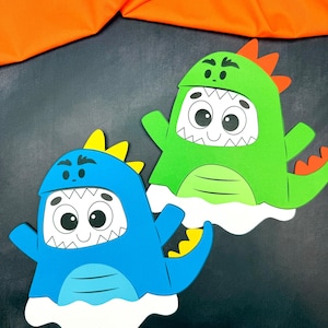 May include: Two colorful dinosaur-themed cutouts. One is blue with yellow accents, the other is green with orange accents. Both have large eyes, white teeth, and a white cloud-like base. The background is dark gray with a bright orange fabric.