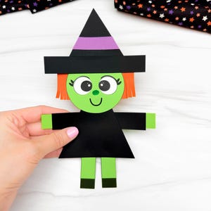 Può includere: Una figura di strega in carta con un viso circolare verde, capelli arancioni e un cappello a punta nero con una fascia viola. La figura indossa un vestito nero con braccia e gambe verdi e scarpe nere. Il viso ha grandi occhi bianchi.