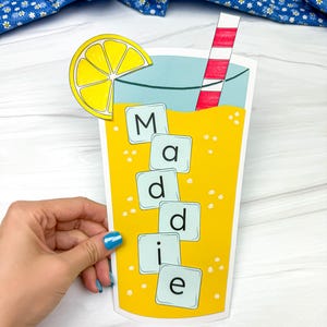 May include: A paper cutout of a lemonade drink with a lemon slice and a red and white striped straw. The word "Maddie" is spelled out in individual letter tiles on the glass. The drink is yellow with white bubbles.