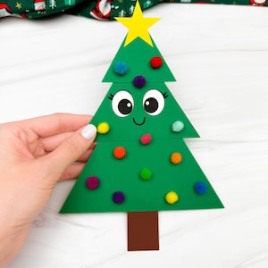 May include: A green Christmas tree decoration with a yellow star on top and colorful pom-poms. The tree has a smiling face with large eyes. A hand holds the tree against a white background.
