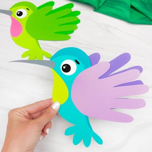 Könnte beinhalten: Zwei bunte Kolibri-Ausschnitte. Einer ist grün mit rosa und der andere blau mit lila Flügeln. Die Vögel haben große Augen und graue Schnäbel. Der Ausschnitt wird von einer Hand gehalten.