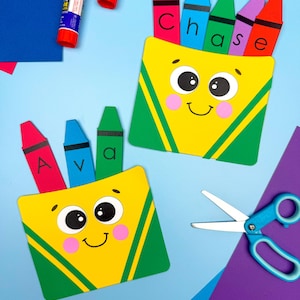 May include: Two yellow crayon box crafts with green accents and smiling faces, each holding colorful crayon cutouts. One box has the name "Ava" and the other has the name "Chase" written on the crayons. Glue sticks and scissors are also visible.