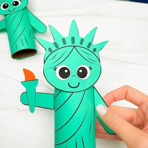 May include: A green paper craft of the Statue of Liberty, complete with a crown, facial details, and a torch. The design mimics the famous monument, with black outlines and a friendly face. The craft is handmade.