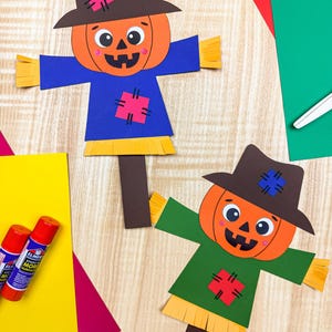 Puede incluir: Dos espantapájaros de papel con cabezas de calabaza naranja, sombreros marrones y camisas azules o verdes. Cada espantapájaros presenta mangas y dobladillos con flecos amarillos, mejillas rosadas y parches coloridos en el sombrero y la camisa.