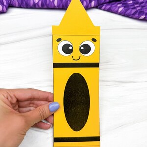 May include: A yellow paper craft shaped like a crayon with a pointed top, a smiling face with large black and white eyes, and a large black oval in the centre. The bottom of the craft features the text simple everyday mom in black letters.