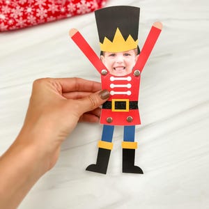 May include: A handmade paper craft of a toy soldier with a child's face. The soldier has a black hat with a gold crown, red jacket, blue pants, and black boots. The arms are raised, and the craft is held by a hand.