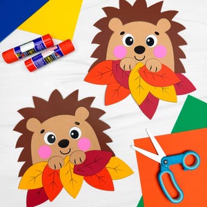 Könnte beinhalten: Zwei Igel-Bastelarbeiten aus Papier mit braunen Stacheln, beigen Gesichtern, rosa Wangen und schwarzen Augen, die jeweils ein Bündel aus roten, orangen und gelben Papierblättern halten, dazu zwei Klebestifte und eine Schere mit blauem Griff.