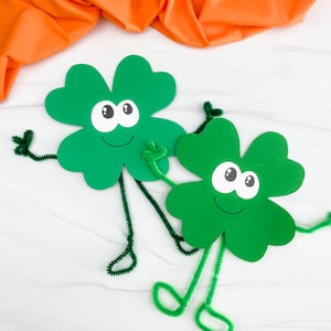 Könnte beinhalten: Zwei grüne Kleeblätter aus Papier mit Wackelaugen, lächelnden Gesichtern und Pfeifenputzerarmen und -beinen. Die Kleeblätter sind vor einem weißen Hintergrund mit einem orangefarbenen Stoffhintergrund platziert und schaffen ein festliches St. Patrick's Day-Thema.