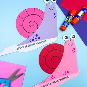 Op de afbeelding: Twee kleurrijke papieren slakkenknutsels met wiebelogen en de tekst "Shell we be friends, Valentine?" worden weergegeven op een blauwe achtergrond. Eén slak is roze met een rode schelp, de andere is paars met een roze schelp. Lijmstiften en scharen zijn ook aanwezig.
