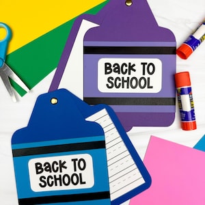 Peut inclure: Deux créations en papier en forme de crayons violet et bleu, chacune avec une étiquette blanche portant le texte noir "BACK TO SCHOOL". La création en forme de crayon bleu est ouverte, révélant du papier ligné blanc. Attache dorée en haut.
