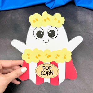May include: A white ghost-shaped cutout with a popcorn theme. The ghost has large black eyes, a smiling mouth, and yellow popcorn around its head. The body is designed as a popcorn container with red and white stripes and the words "POP CORN".