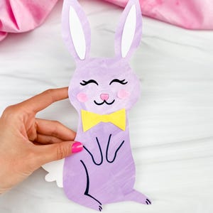 May include: A lilac paper bunny with white inner ears, pink cheeks, and a yellow bow tie. The bunny has black whiskers and eyelashes. The bunny is a craft project, possibly for Easter or spring decorations.