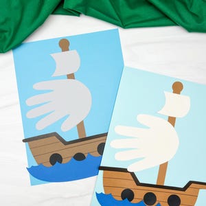 May include: Two handmade paper crafts of ships on a light blue background. The ships are brown with white sails shaped like a handprint. The waves are blue, and the masts are brown. The background is white with a green cloth.