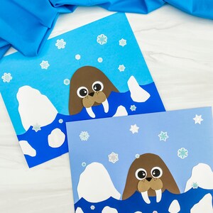 May include: Two handmade cards featuring a cartoon walrus design. The walruses are brown with large eyes and tusks, emerging from blue water. White iceberg shapes and snowflake stickers decorate the light and dark blue backgrounds. A blue fabric is draped in the background.