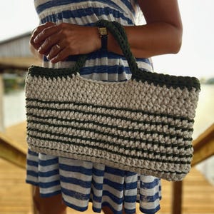 Puede incluir: Un bolso tote de crochet hecho a mano con cuerpo color crema y ribetes y asas verde oscuro. El bolso presenta rayas horizontales verde oscuro. El bolso lo sostiene una persona que lleva un vestido a rayas azules y blancas.