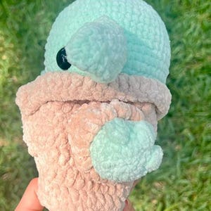 May include: A handmade crochet plush toy in the shape of a character with a large head and ears. The toy features a light blue head and ears, a tan body, and a black eye. The plush is made of soft, textured yarn.