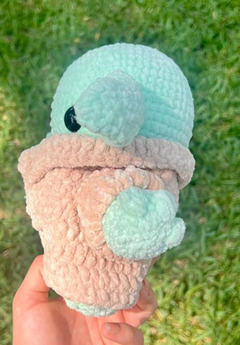 May include: A handmade crochet plush toy in the shape of a character with a large head and ears. The toy features a light blue head and ears, a tan body, and a black eye. The plush is made of soft, textured yarn.