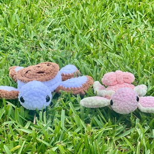 May include: Two crocheted turtle plushies rest on green grass. One turtle is white, brown, and tan, resembling a cookie. The other is pink and light green. Both have black eyes and are handmade.