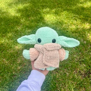 May include: A crocheted doll in the likeness of a popular character. The doll has large ears, black eyes, and is primarily mint green and tan. The doll is held in a person's hand against a background of green grass.