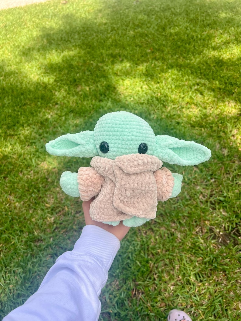May include: A crocheted doll in the likeness of a popular character. The doll has large ears, black eyes, and is primarily mint green and tan. The doll is held in a person's hand against a background of green grass.