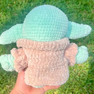 May include: A handmade crochet doll in the likeness of a character from a popular science fiction series. The doll features a light teal head and ears, with a tan-colored body and arms. The doll is made of a soft, plush yarn.