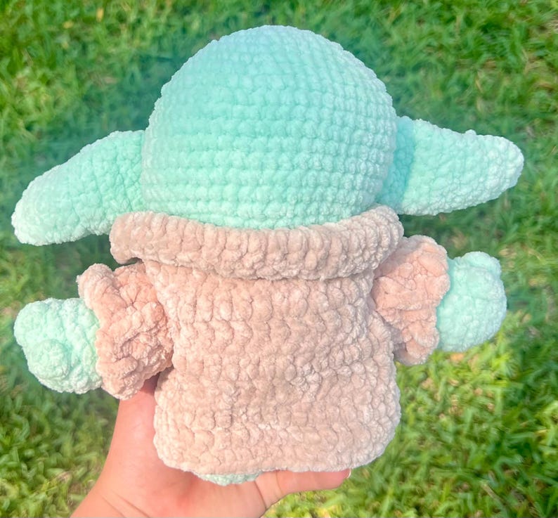 May include: A handmade crochet doll in the likeness of a character from a popular science fiction series. The doll features a light teal head and ears, with a tan-colored body and arms. The doll is made of a soft, plush yarn.