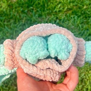 May include: A handmade plush toy in the shape of a baby, held in a hand. The toy features a light pink and mint green color scheme. The plush has a soft, textured appearance, with the head and arms in mint green and the body in light pink.
