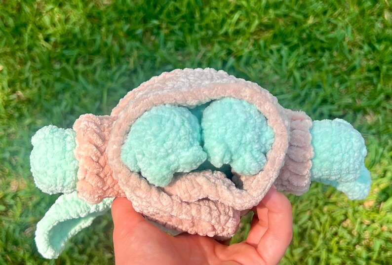 May include: A handmade plush toy in the shape of a baby, held in a hand. The toy features a light pink and mint green color scheme. The plush has a soft, textured appearance, with the head and arms in mint green and the body in light pink.