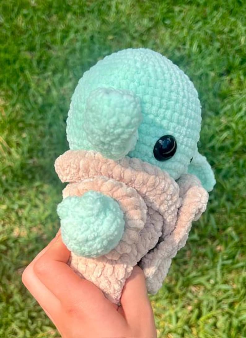 May include: A handmade crochet octopus in light teal and beige. The octopus has a round head, two large black eyes, and soft, textured tentacles. The plush toy is held against a background of green grass.